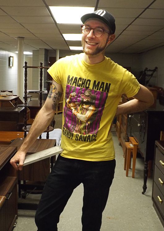 A smiling man in a yellow graphic T-shirt & black cap stands in the church basement preparing for the Tag Sale.