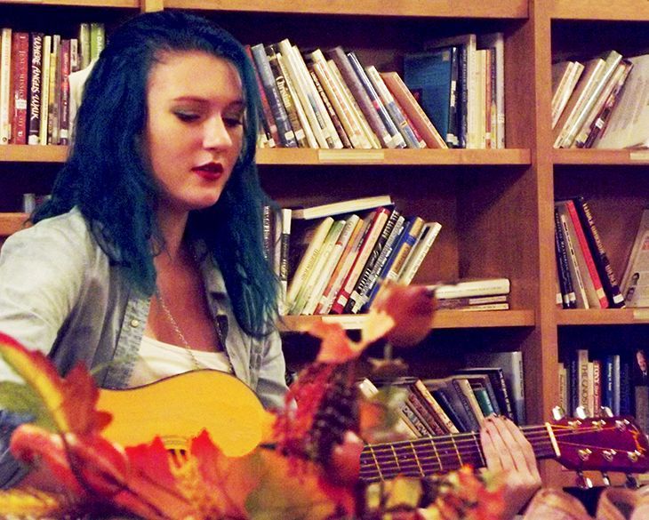 A blue-haired young woman plays acoustic guitar in front of a wall of full bookshelves