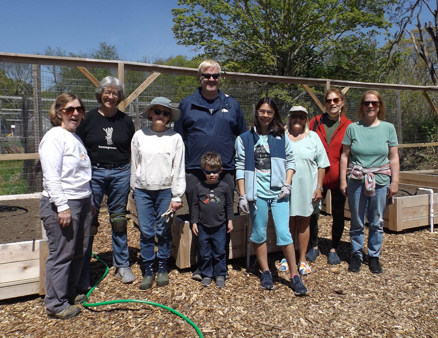 A group of people stands outdoors with Pastor Charlie in the Teaching Garden, beside raised planter boxes near a fence