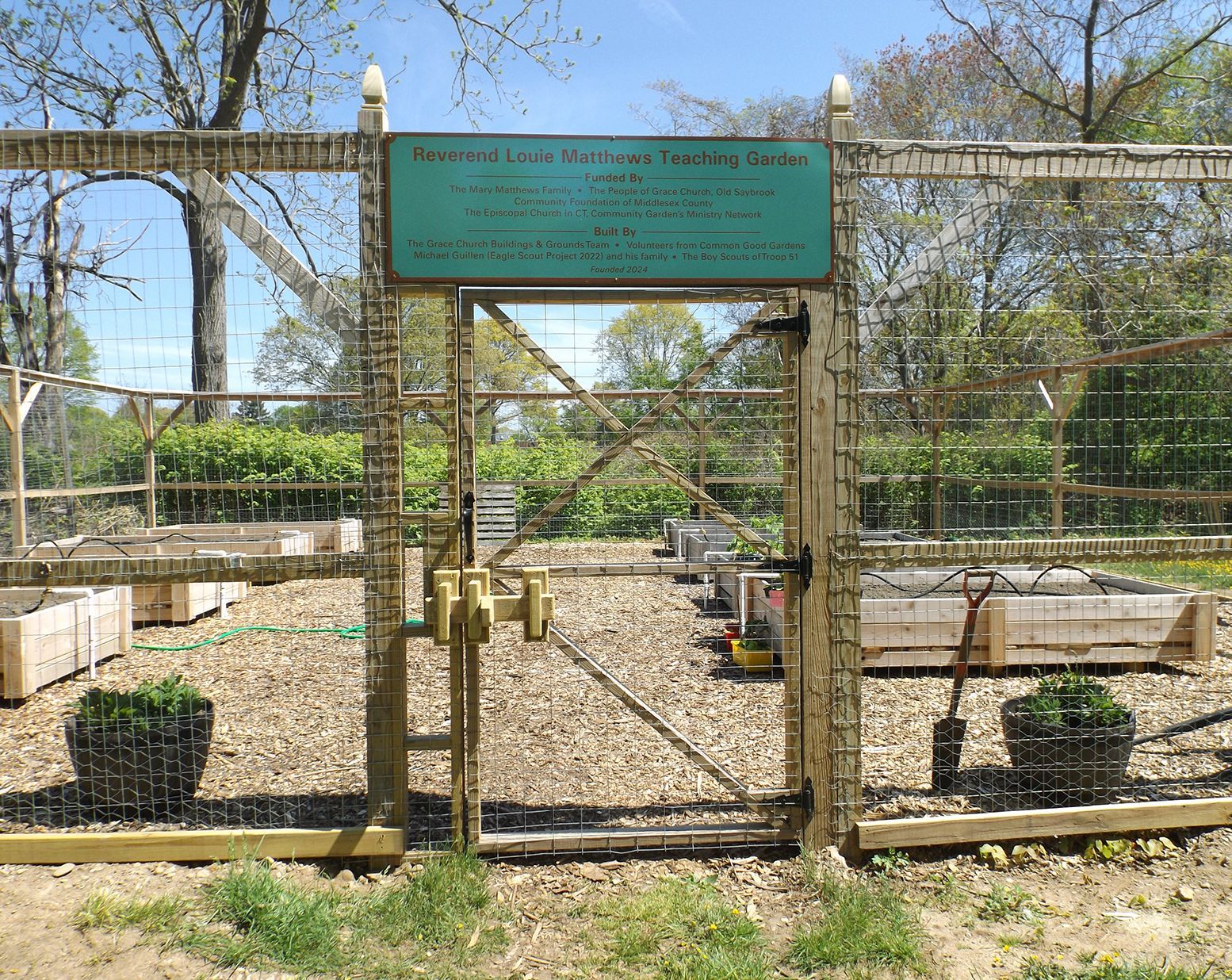 Fenced community teaching garden with raised beds & potted plants under a green sign.