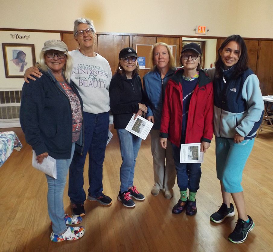 Group of 6 people standing in the Parish Hall, smiling & holding papers, w/framed artwork on the wall behind them.