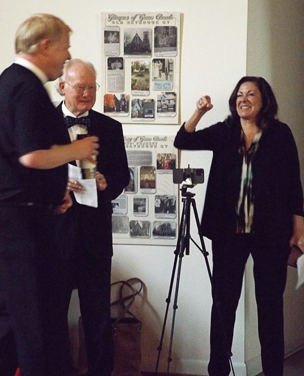 The Pastor chats with a small group indoors beside a poster display & camera tripod; while a woman raises a fist & smiles.