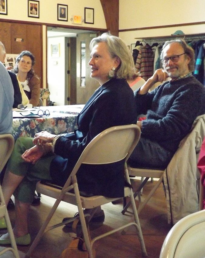 People seated in folding chairs around a table chuckle while listening to a speaker