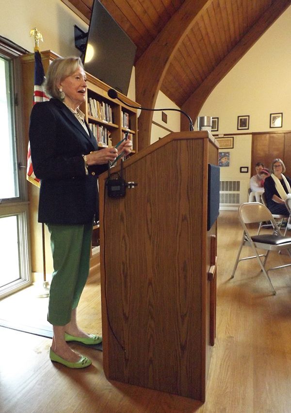 A woman in a navy suit jacket speaks at a wooden podium in the Parish Hall w/bookshelves & seated attendees nearby