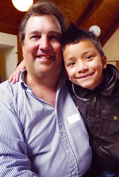 Smiling man and child lean together in the Parish Hall, with wooden ceiling and white wall background.