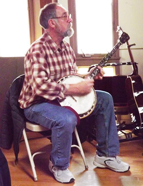 A bearded man wearing a plaid shirt and jeans while seated in the Parish Hall, plays banjo near a window
