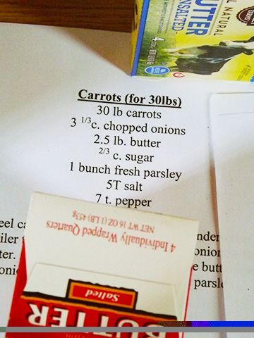 A recipe card listing ingredients for a large group serving of carrots, beside packages of butter on a countertop.