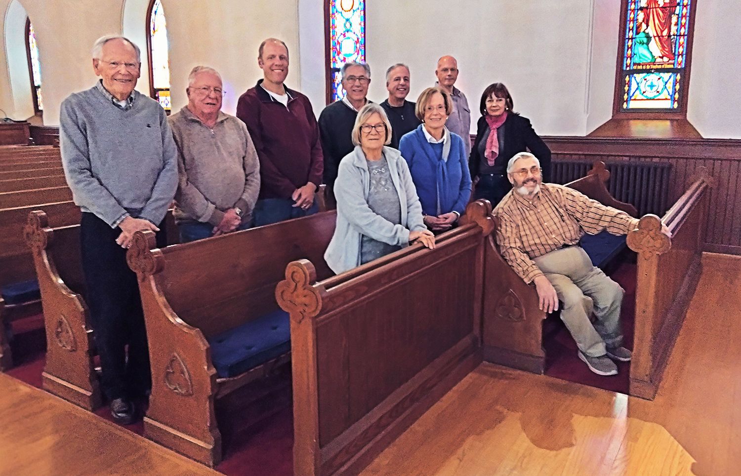 Group of Ushers posing together in wooden church pews, with stained-glass windows in the background.