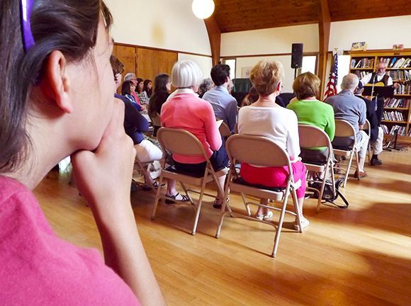 Audience seated in folding chairs in a hall, listening to a speaker.