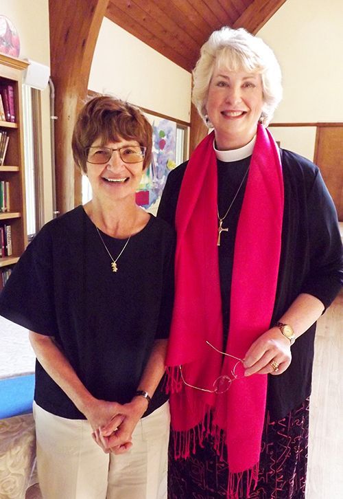 Two smiling women stand together in Grace Church's Parish Hall, beside some bookshelves.