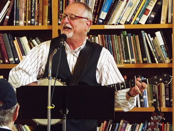 Man sings into a microphone in front of the Parish Hall bookshelves, holding a banjo.