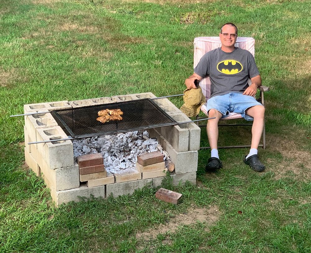 Man wearing a Batman T-shirt sits in a lawn chair beside a cinder block fire pit in the grassy churchyard.