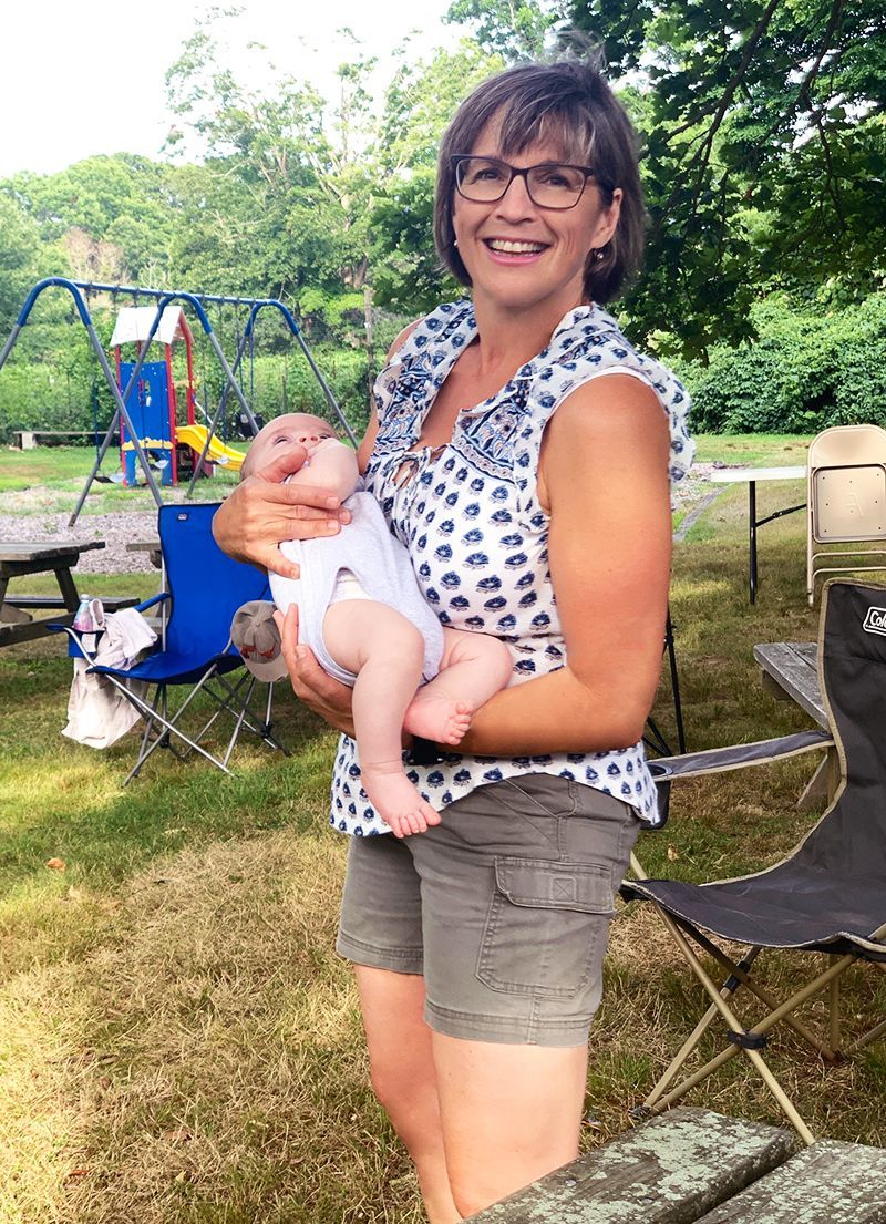 A smiling woman in a blue-patterned blouse holds a baby outdoors in the churchyard w/playground equipment in the background