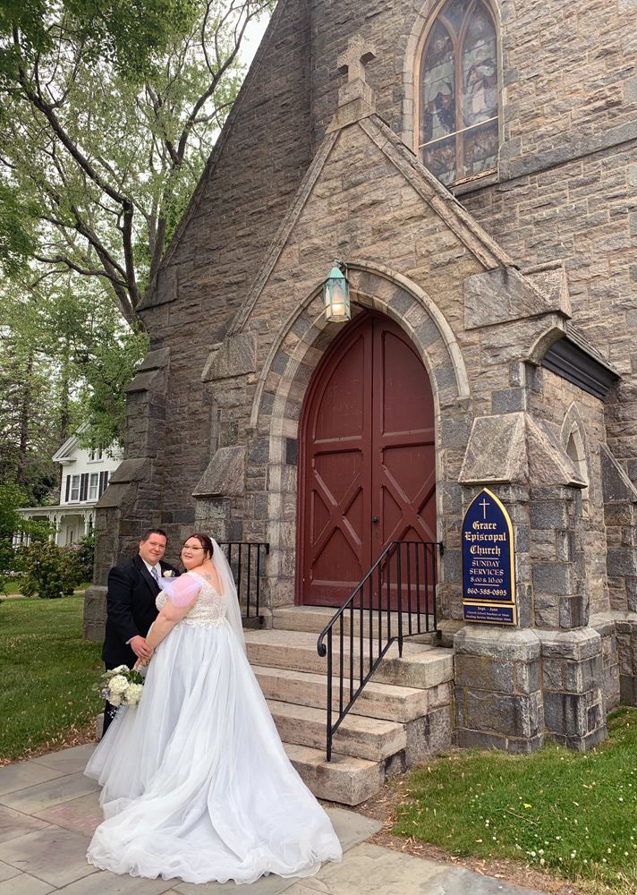 A bride and groom pose outside the stone church entrance, bride in a flowing white gown and groom in a black tuxedo