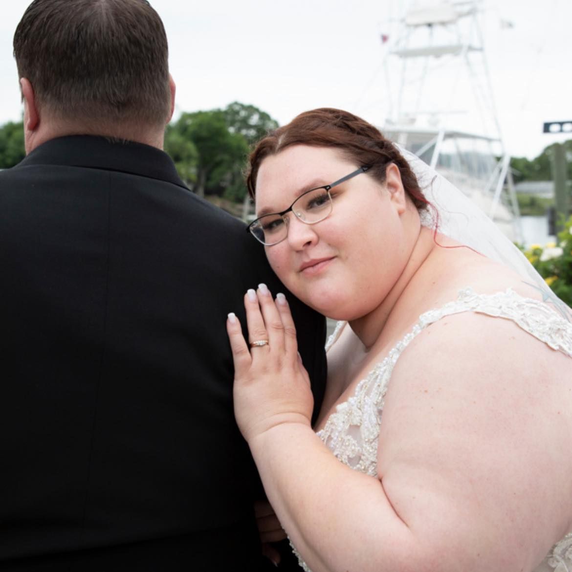 A bride rests her head on her new husband's back, while standing outdoors in her gown & veil, displaying her wedding ring
