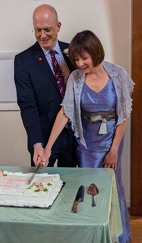A newly married husband & wife cut a frosted sheet cake together, at a table covered with a green cloth.