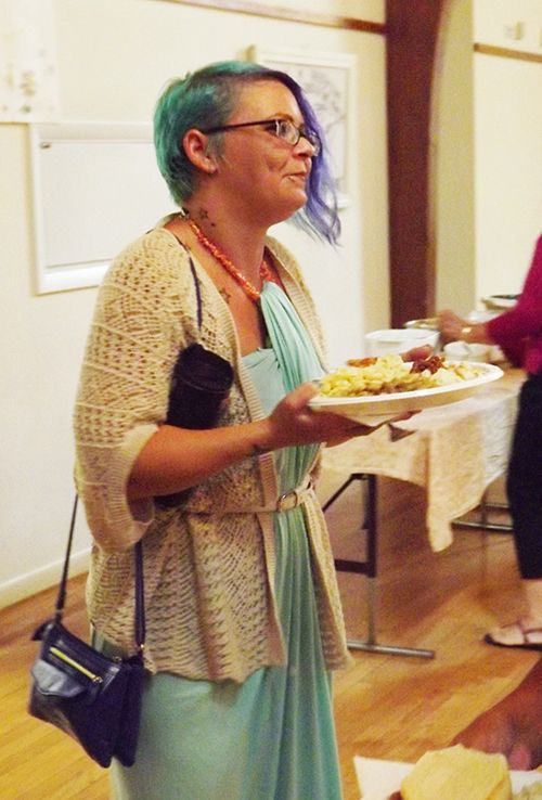 A woman with festive multicolor hair stands in a turquoise dress and beige cardigan, with a plate of food at an indoor event.
