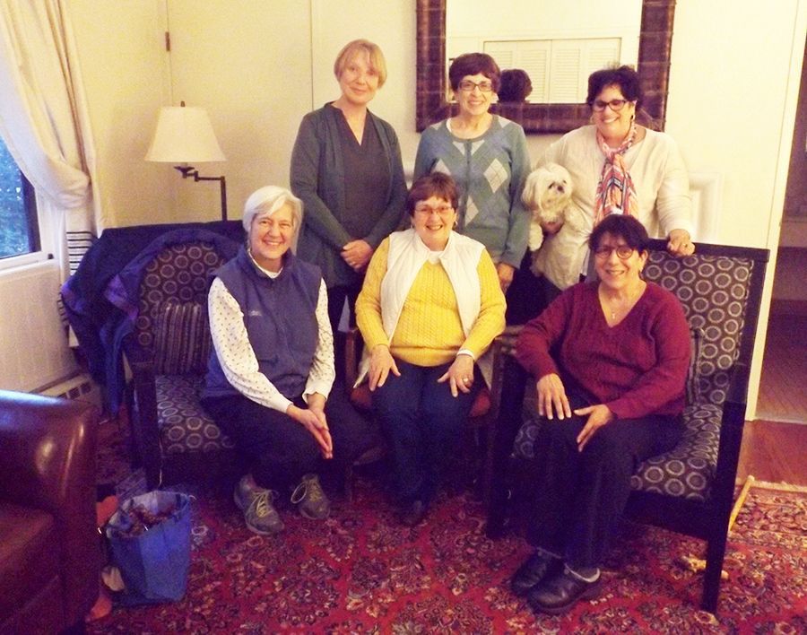 6 women & a small dog pose in the cozy Hudson Room w/a lamp, couch, mirror, patterned chairs, & a red rug