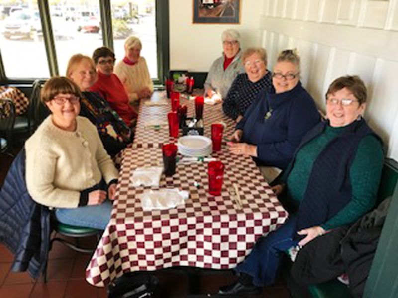 A group of friendly women sit together around a red-checkered table in a restaurant dining-room preparing for a meal together