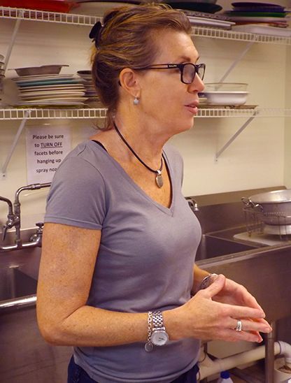 A woman stands in profile in a kitchen, near a sink and shelves, speaking w/her hands folded.