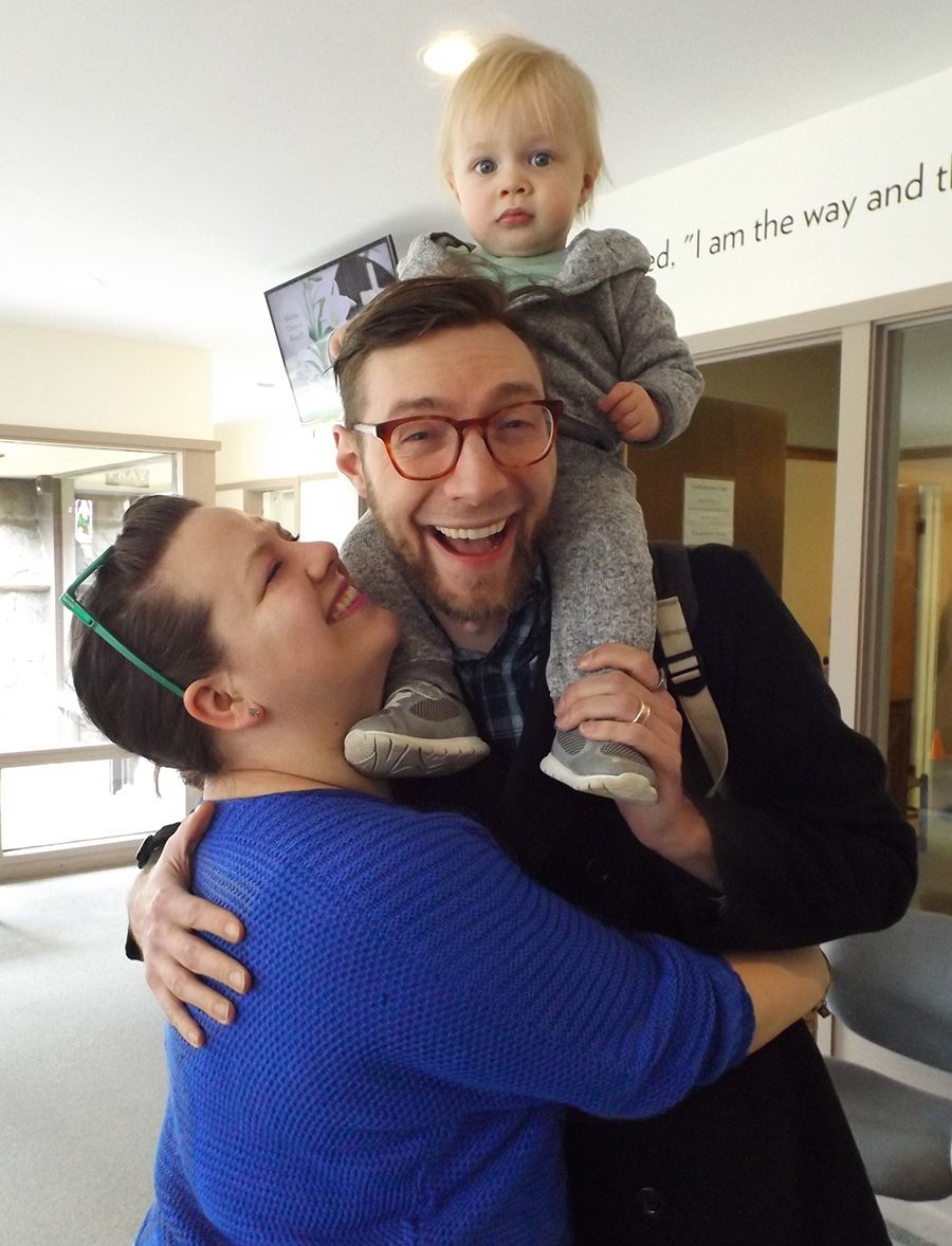 Parents laughing & hugging, while the father holds their toddler son on his shoulders.