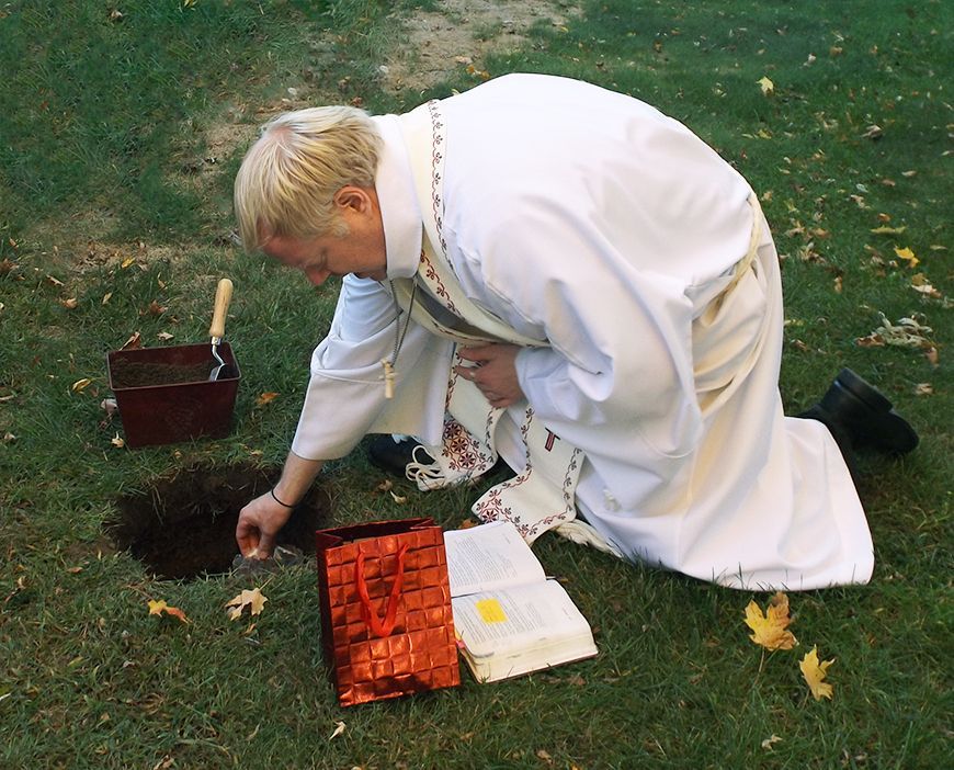 The Pastor kneels in white robes outdoors, placing soil & ashes into a grave during a burial in the church's Memorial Garden