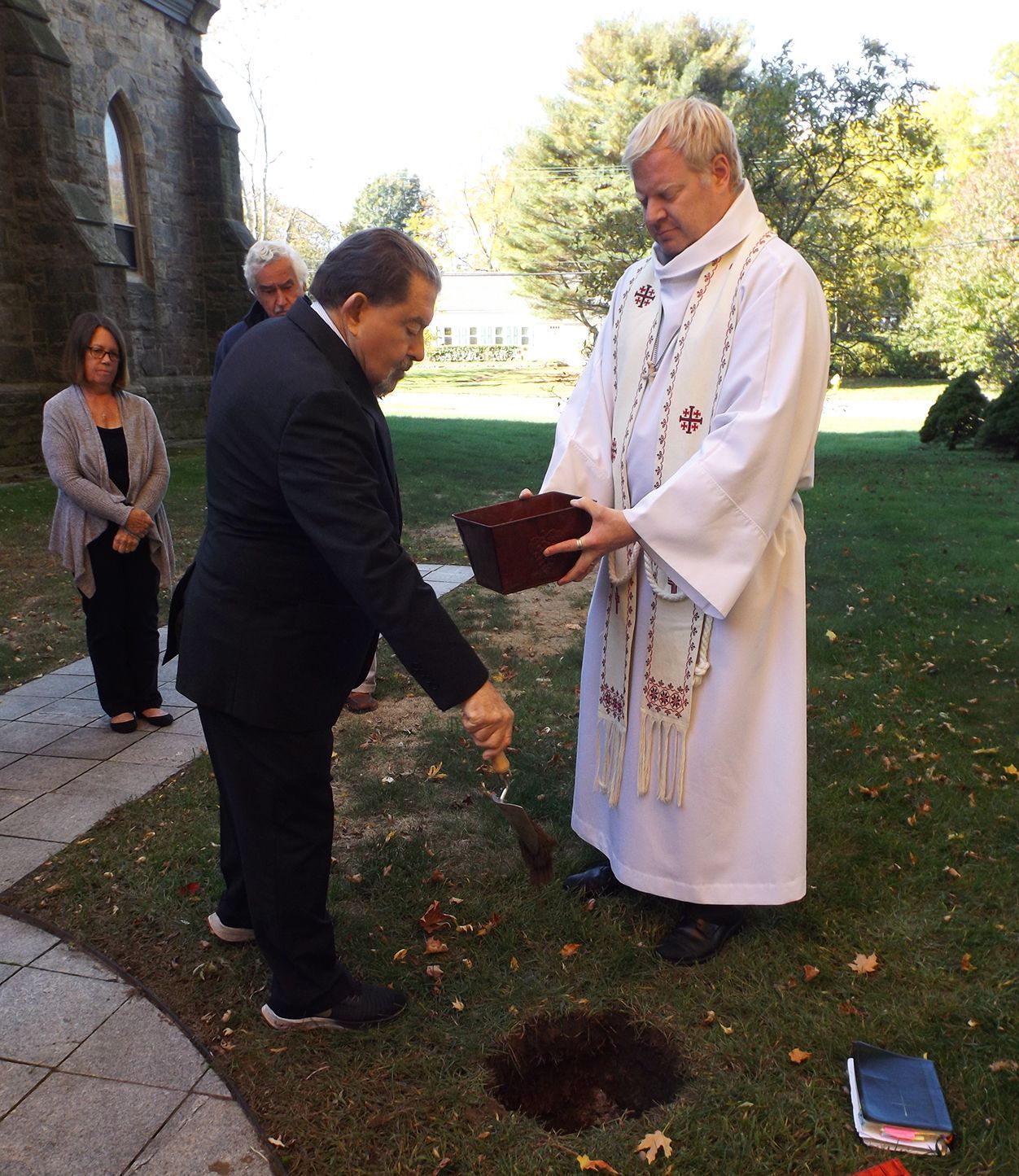 A man empties a small shovelful of dirt into a burial hole during a ceremony in the Memorial Garden w/the Pastor standing by