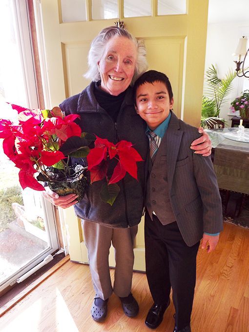 A smiling elderly woman stands at her open front door with her arm around a young boy delivering her a red poinsettia plant