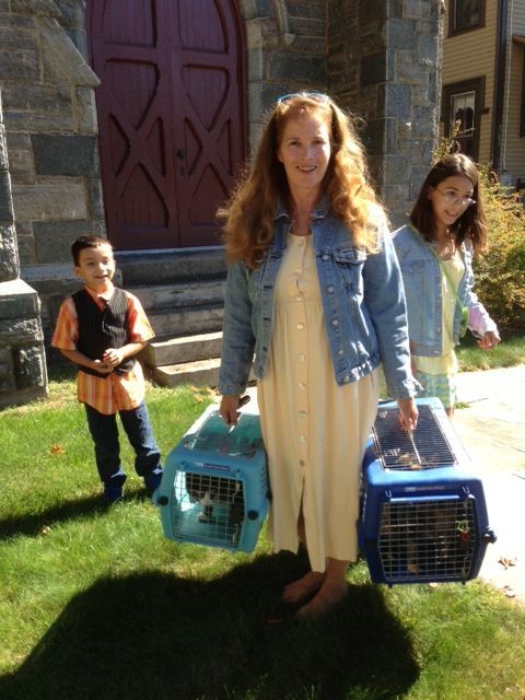 An auburn-haired woman in yellow dress w/denim jacket carries 2 pet crates on the grass outside our stone church w/2 children