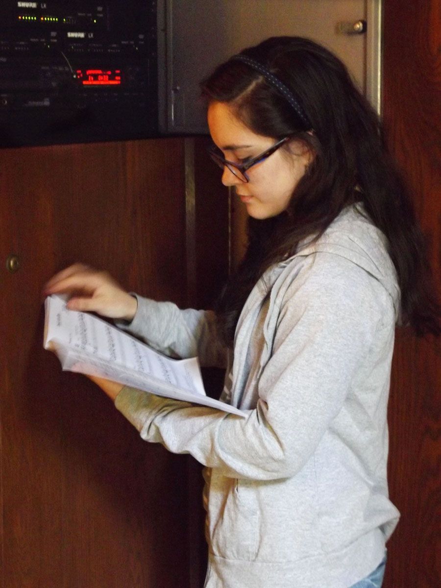 A young woman in glasses looks through a sheaf of papers while standing beside a panel of electronic recording devices