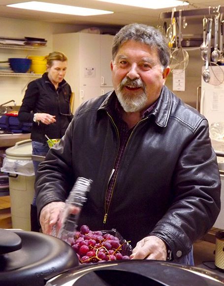 A smiling man wearing a dark leather jacket stands in the kitchen serving grapes, with a woman working in the background.