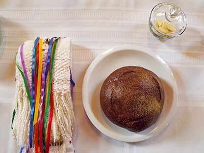 A small round loaf of dark brown bread, sits on a white tablecloth near a glass jar of wafers & a strap w/colorful ribbons.