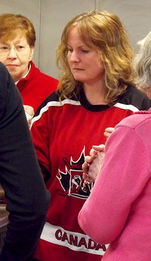 Woman in a red Canada hockey jersey stands indoors among a group, looking down.