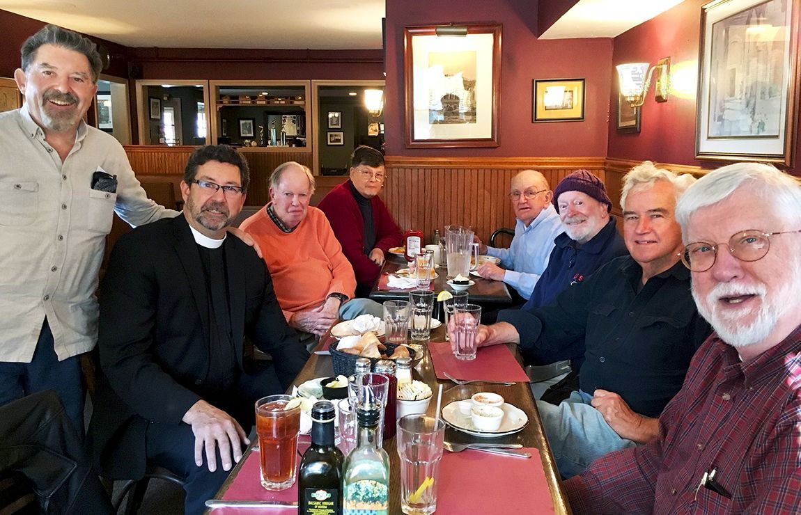A group of men are seated around a restaurant table, smiling & chatting over beverages in a warm dining room