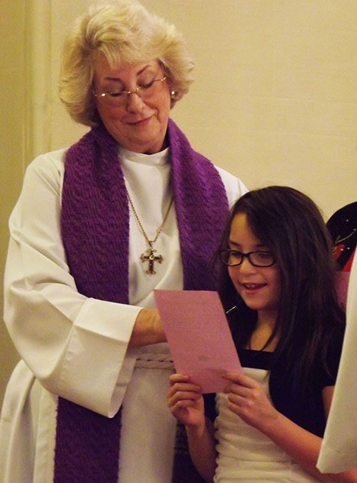 A female pastor in a white robe and purple stole stands with a dark-haired young girl reading a pink paper in church