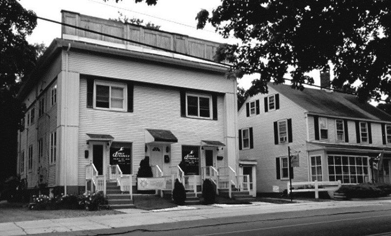 A 2-story house w/porch & shutters stands beside a smaller house on a tree-lined street.