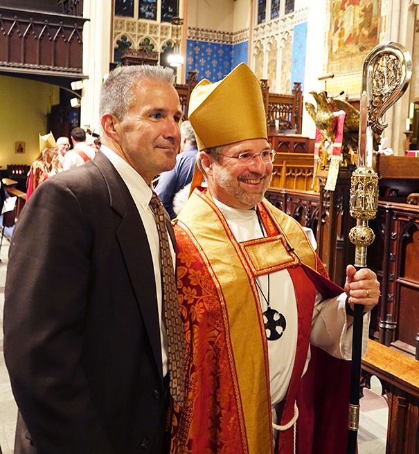 A male parishioner stands in church beside the smiling former Bishop Ian, Douglas who wears ornate vestments & holds a staff