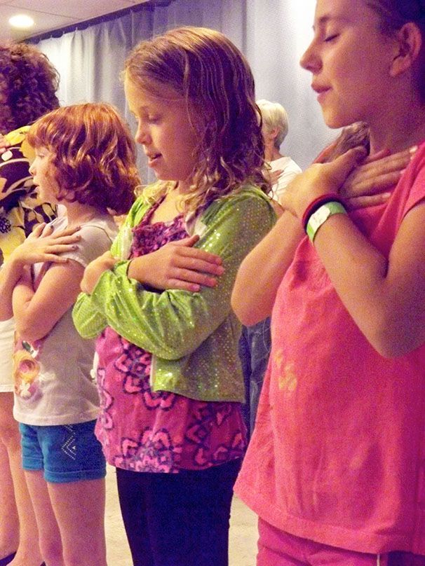 3 young girls stand w/their eyes closed & their hands on their hearts, while singing