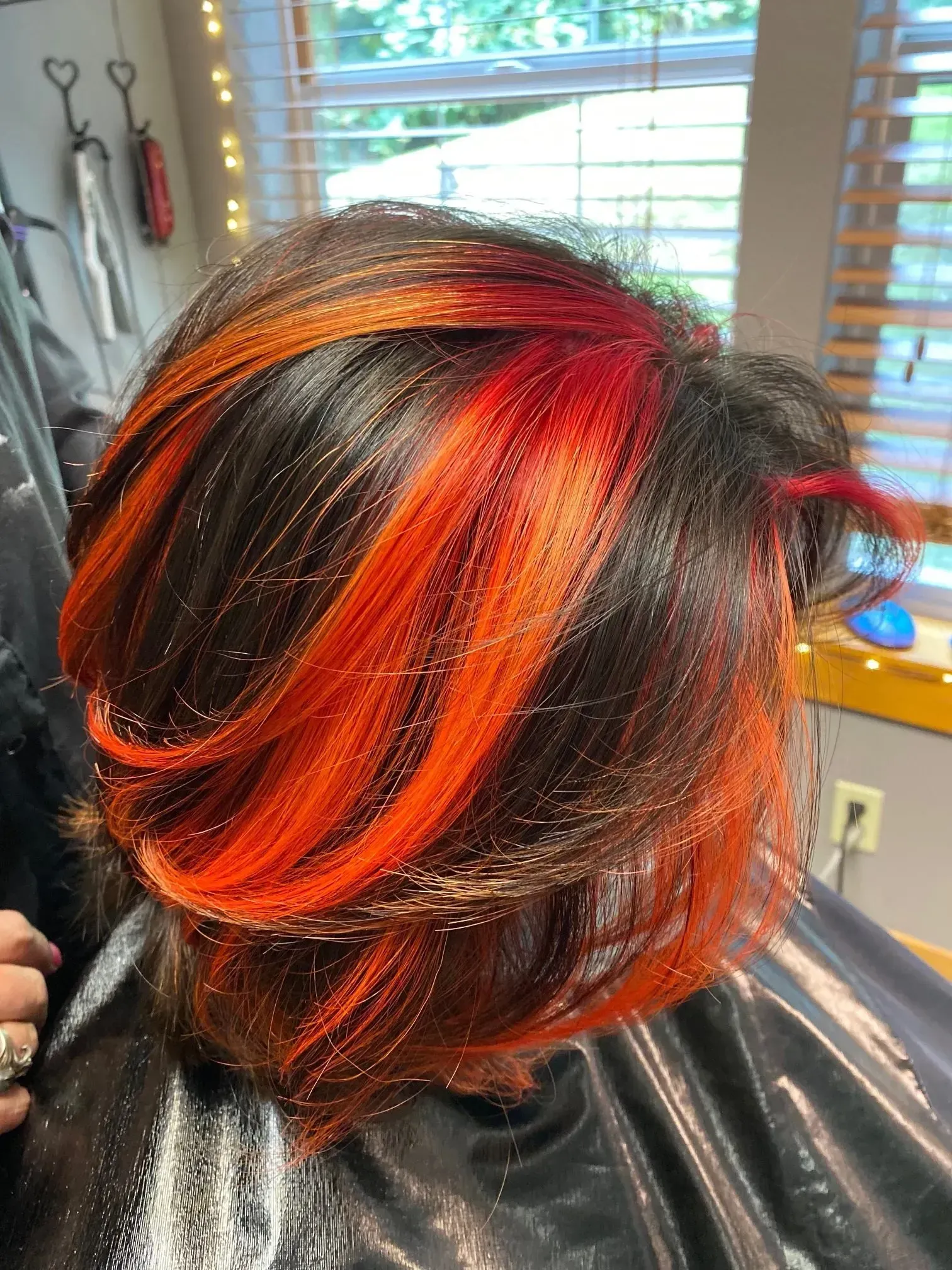 Short dark hair with bright orange-red highlights in a salon chair near a window