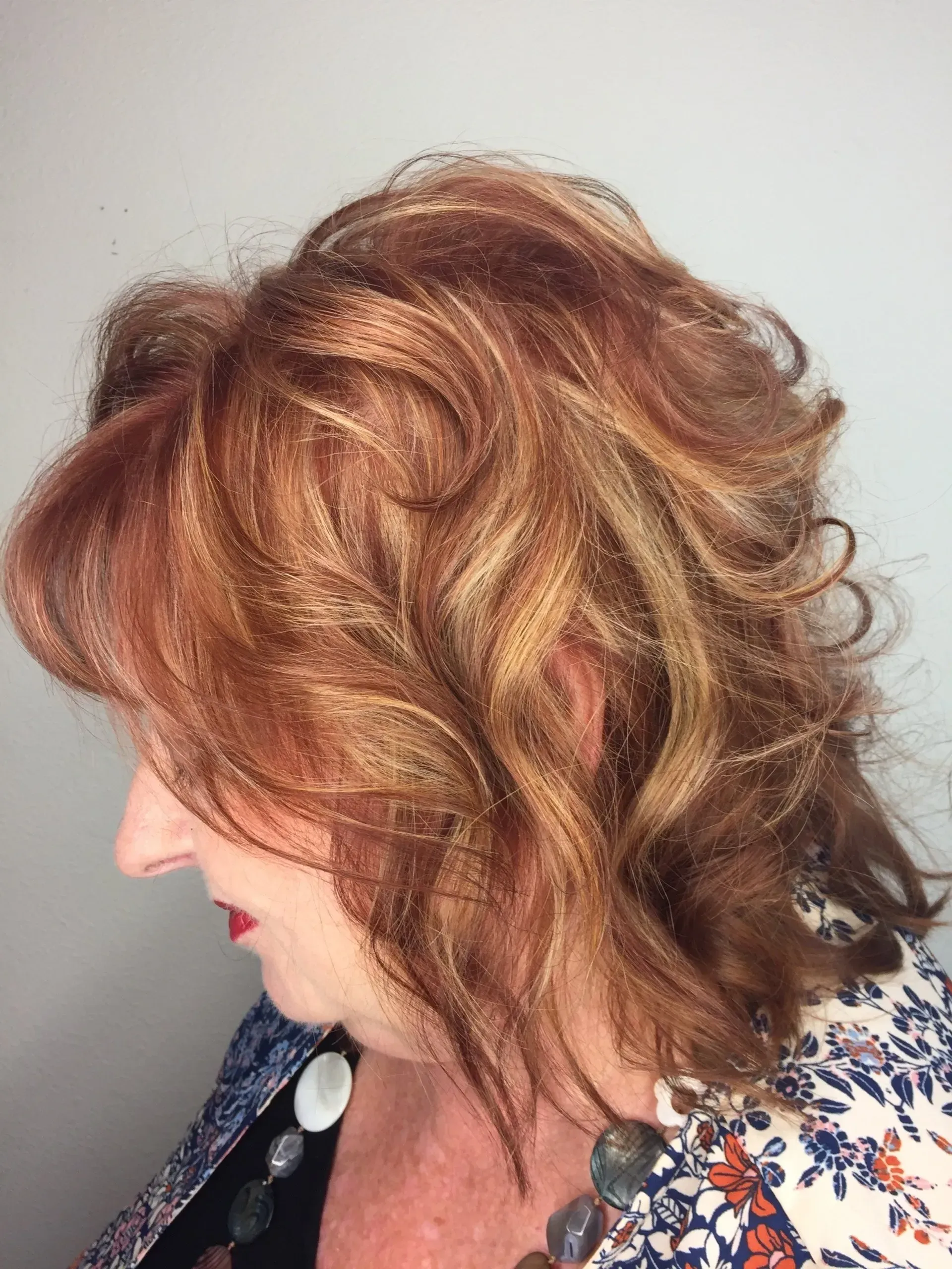 Side-profile of a person with wavy auburn hair wearing a floral top against a plain wall