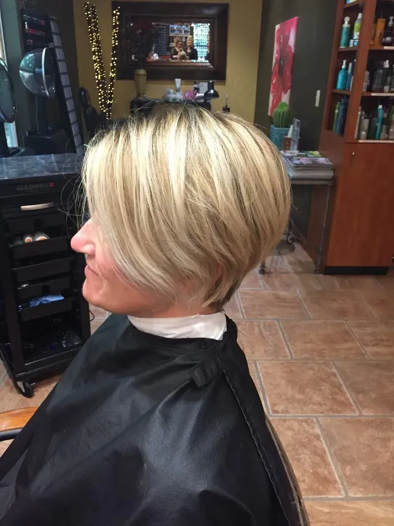 Side-profile of a blonde woman with a short bob haircut in a salon chair, wearing a black cape.