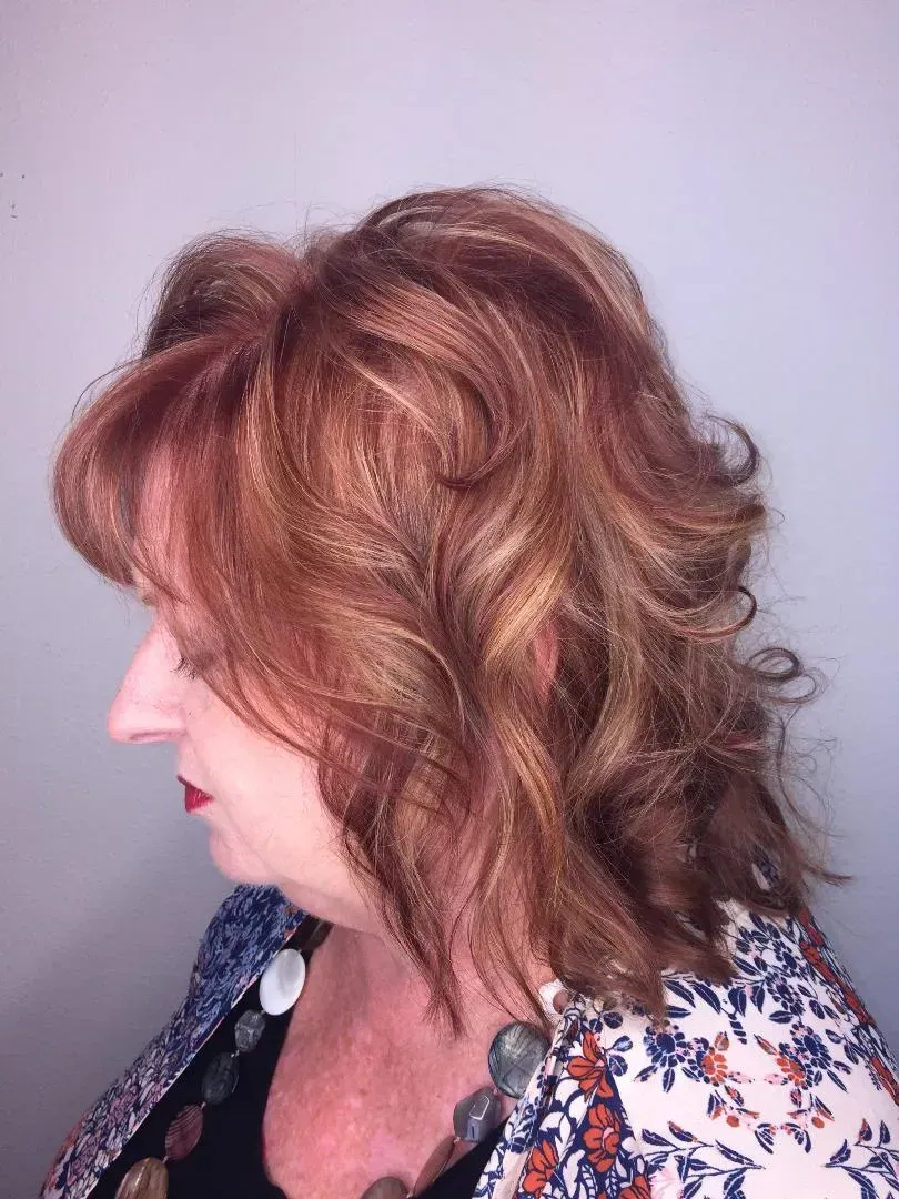 Side-profile portrait of a person with wavy reddish hair, wearing a floral top against a plain wall.