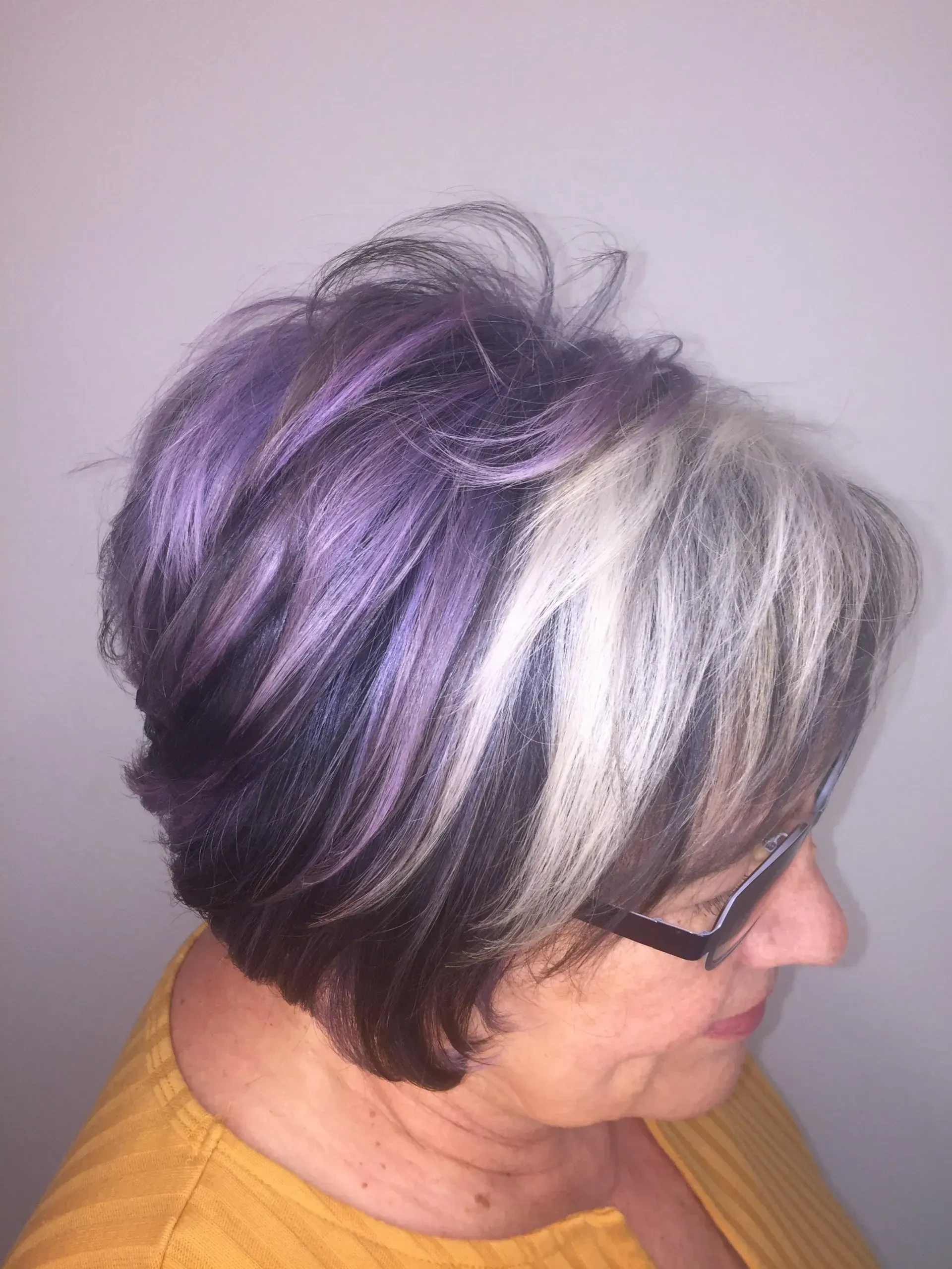 Side-view of a woman with short purple and silver hair, wearing glasses and a mustard sweater.