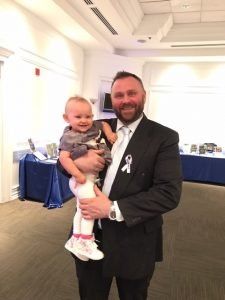 Man in a black suit holding a smiling baby in a gray outfit at an indoor event hall