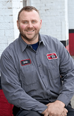 Man in gray work shirt sitting and smiling against a white wall.