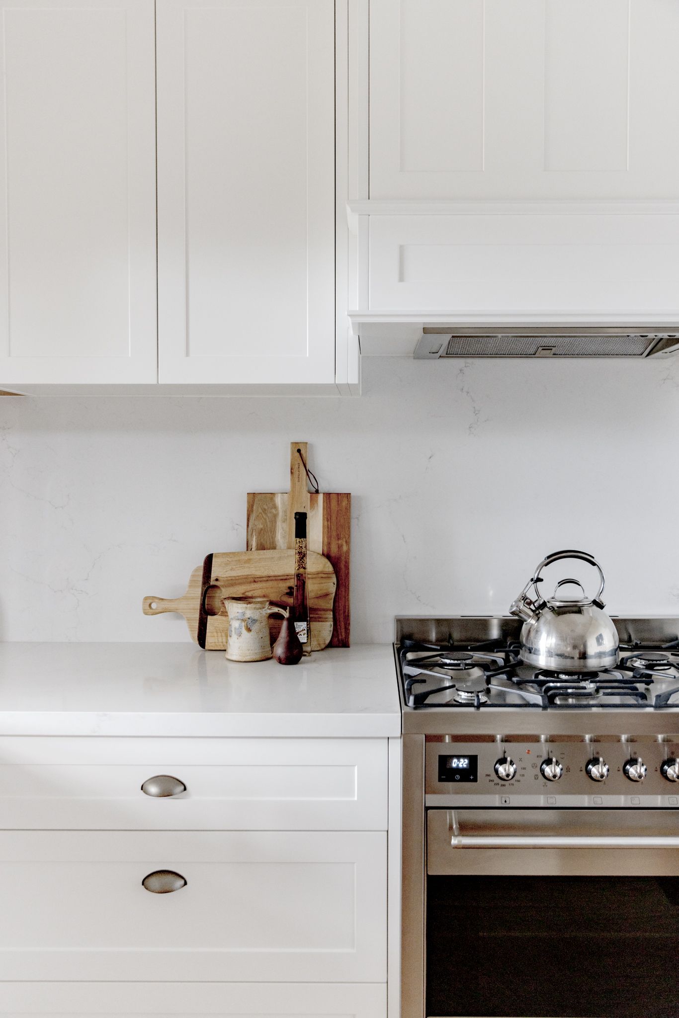 A locally manufactured kitchen in Newcastle NSW, featuring a stove and oven. White shaker style joinery with brushed nickel cup handles and matching white benchtops and stone splash back.