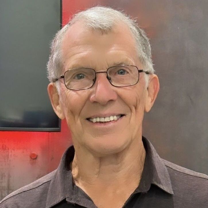 Man with glasses smiles, wearing a dark shirt in front of a red and gray background.