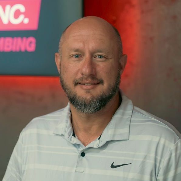Man with a beard wearing a light blue polo shirt with a Nike logo, standing in front of a red sign.
