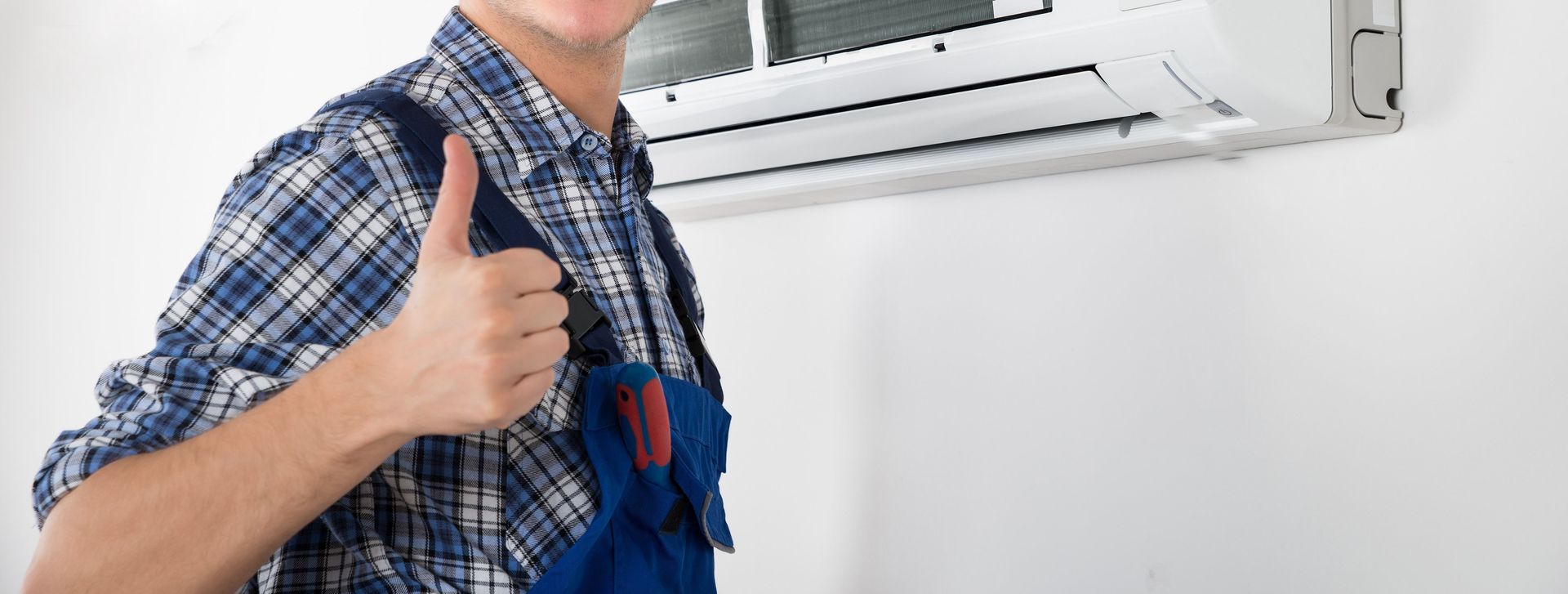 HVAC technician giving thumbs up next to installed air conditioner on white wall.