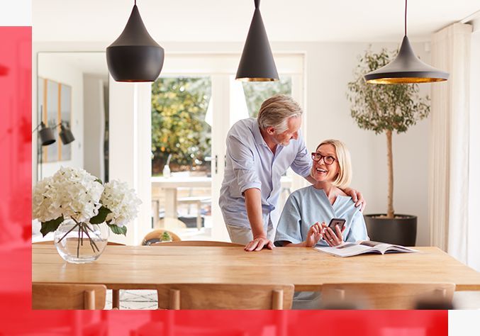 Couple in light-filled dining room, one with arm around the other, looking at a phone.